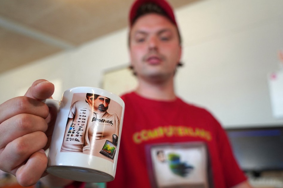 Asker Bryld Staunaes, member of artists’ collective Computer Lars, poses wearing 'Computer Lars' branded t-shirt and cap and holds a mug with an AI-generated image of Marcel Proust in Aarhus, Denmark, on August 3, 2022. James Brooks, AFP